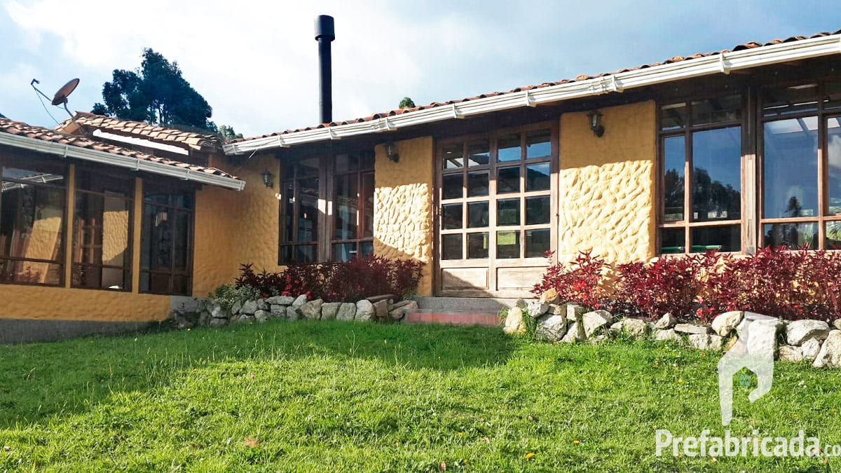 Casa prefabricada campestre con teja de barro, chimenea, ventanas grandes y jardín con flores