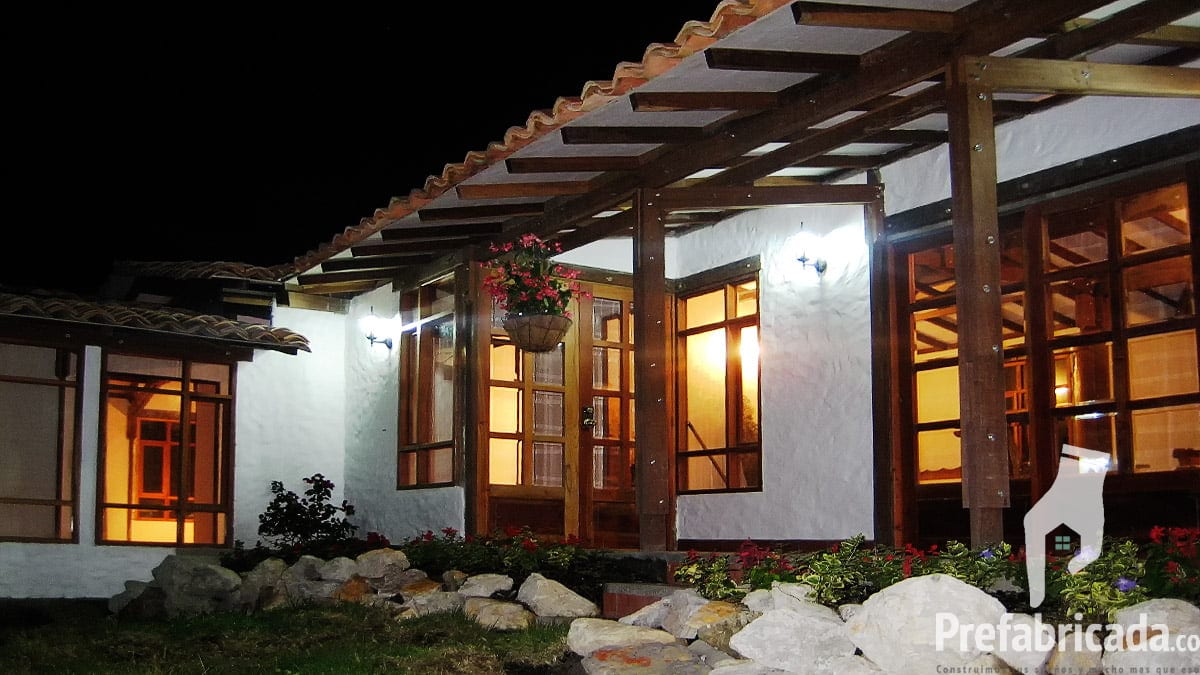 Casa prefabricada campestre con porche de madera iluminada al atardecer y jardín con piedras