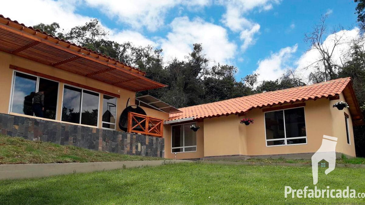 Casa prefabricada de teja de barro color terracota, fachada beige y ventanal amplio hacia el jardín