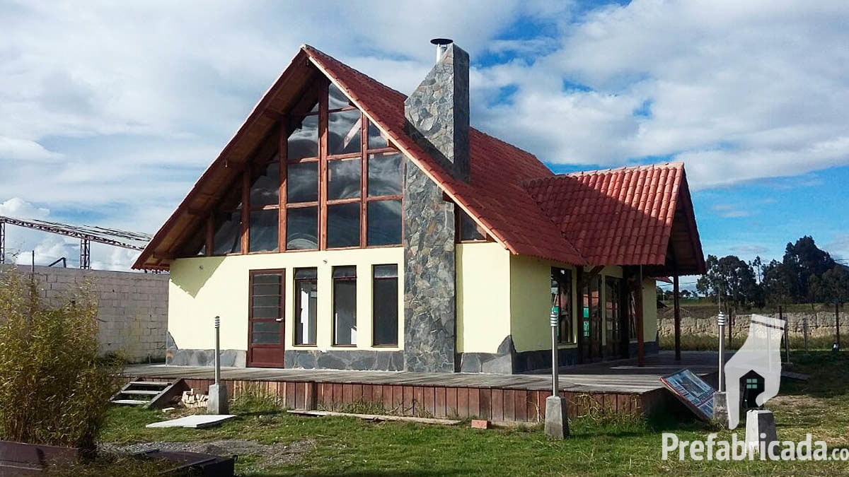 Casa prefabricada con techo a dos aguas rojo, chimenea de piedra y ventanal, rodeada de pinos