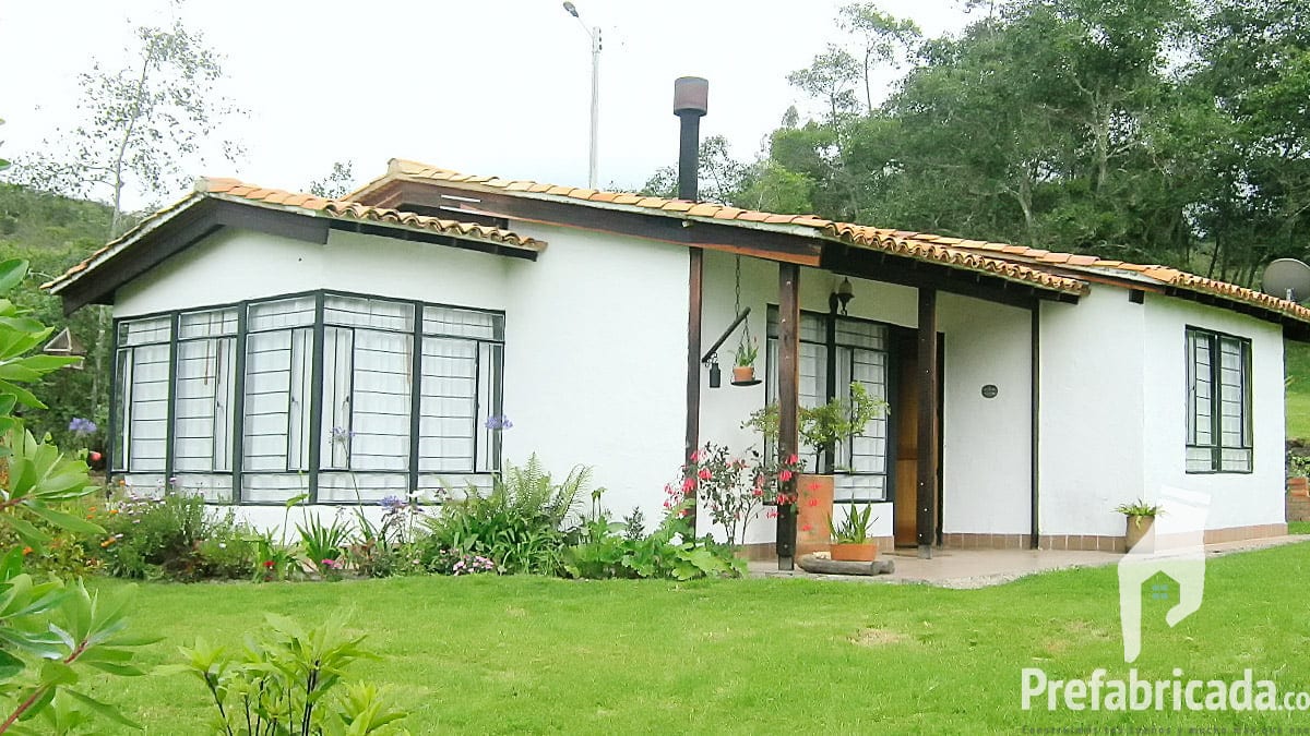 Casa prefabricada blanca de una planta con ventanal de piso a techo, cubierta a un agua y jardín con arbustos