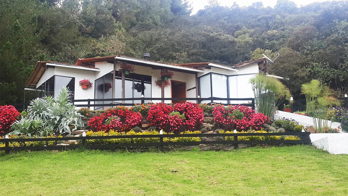 Casa prefabricada blanca con cubierta oscura, jardín con flores rosadas y colinas al fondo