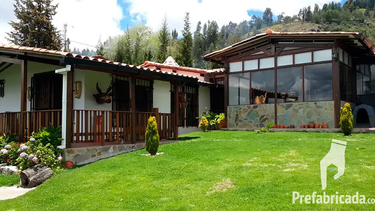 Casa prefabricada con ventanal amplio y porche de madera, rodeada de árboles y jardín