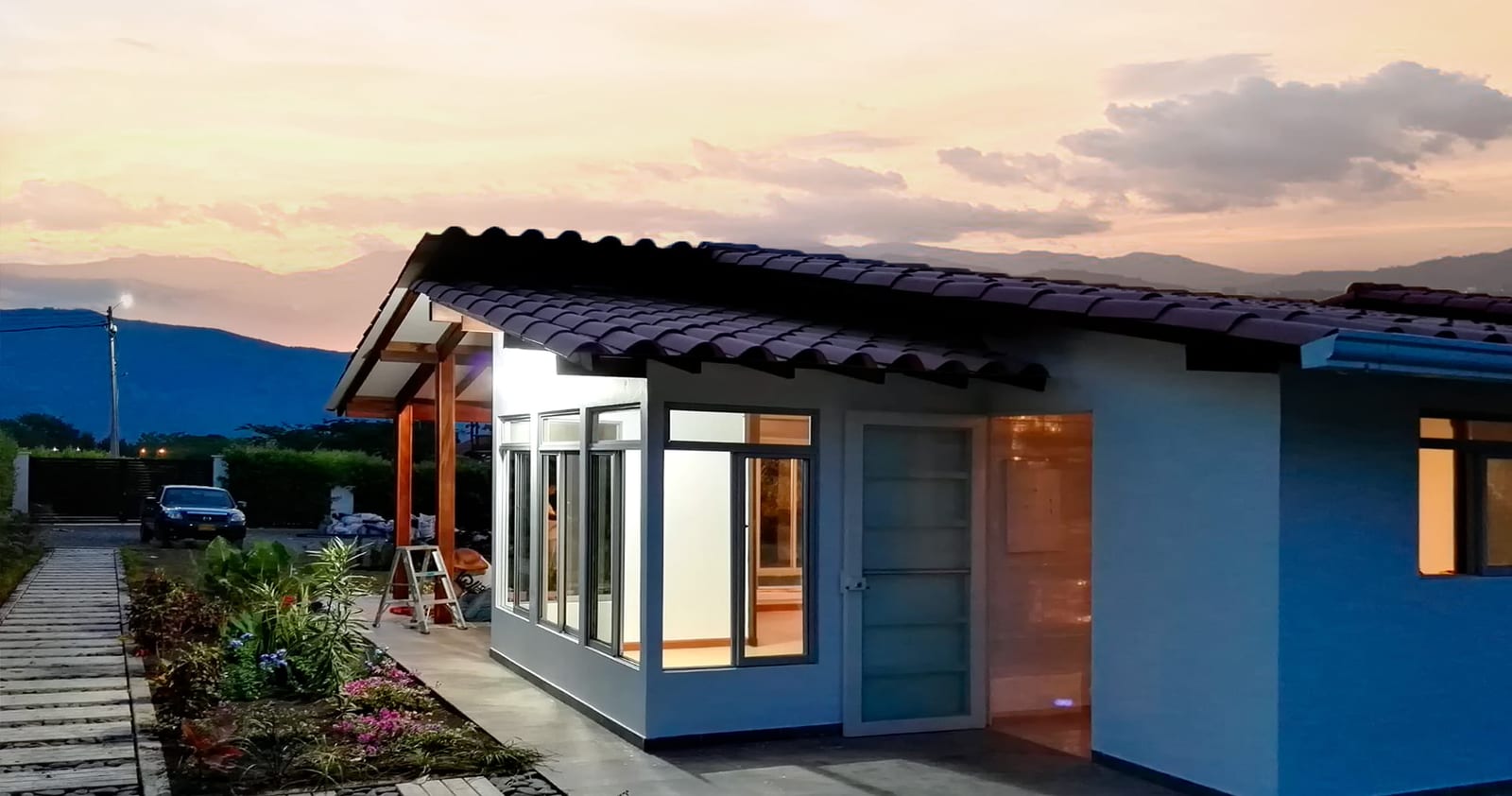 Casa prefabricada moderna con teja oscura y ventanal iluminado al atardecer, con montaña al fondo